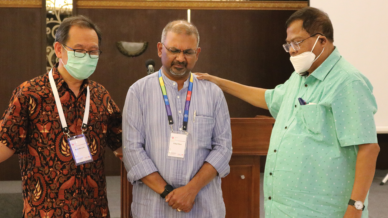 Sunoko Lin, left, and Bijoy Roul, right, pray for Mennonite Church in India executive secretary Vikal Rao at the 2022 Mennonite World Conference assembly in Indonesia. — Mennonite World Conference