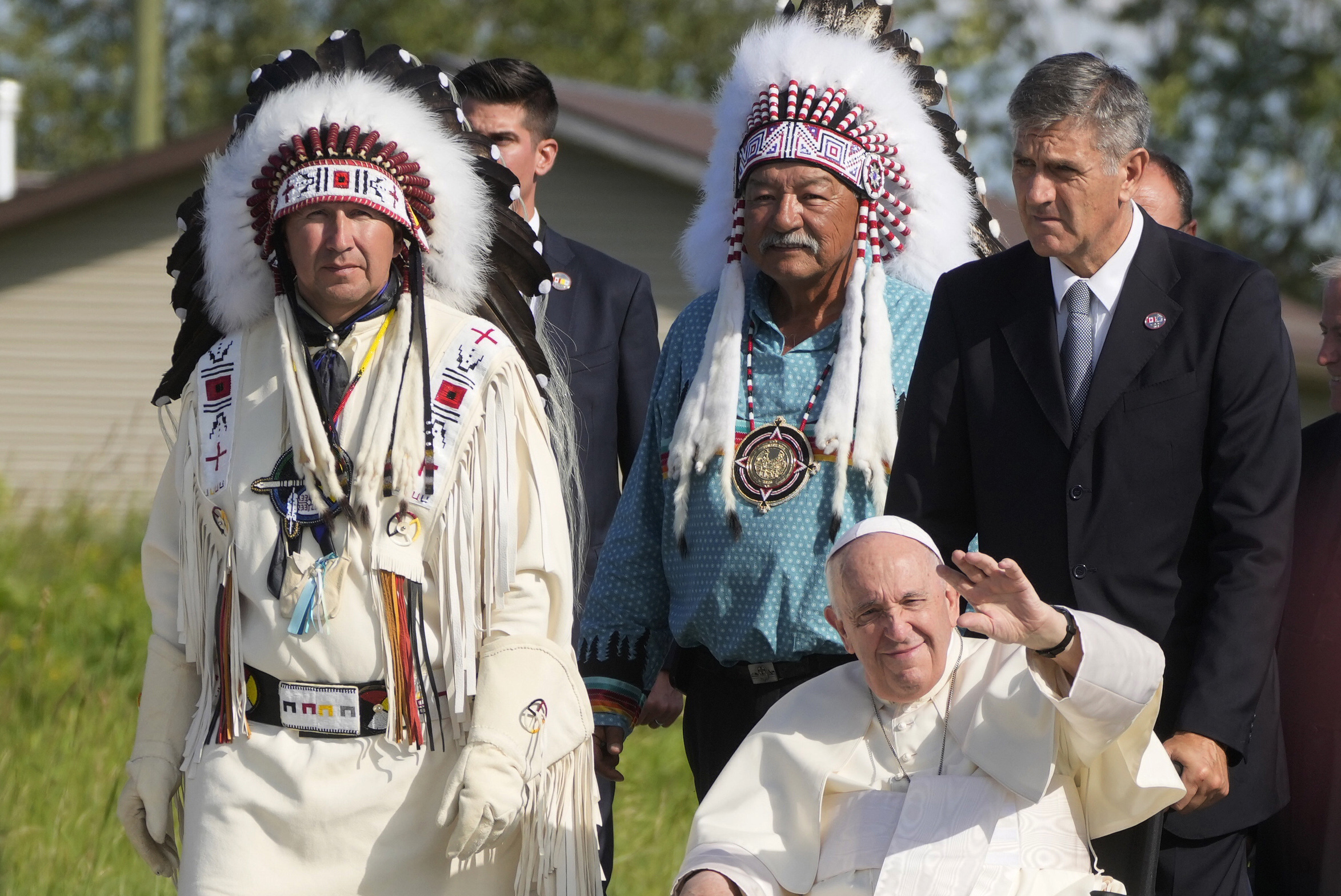 Two Indigenous men in regalia stand to the left of Pope Francis, who sits in a wheel chair.