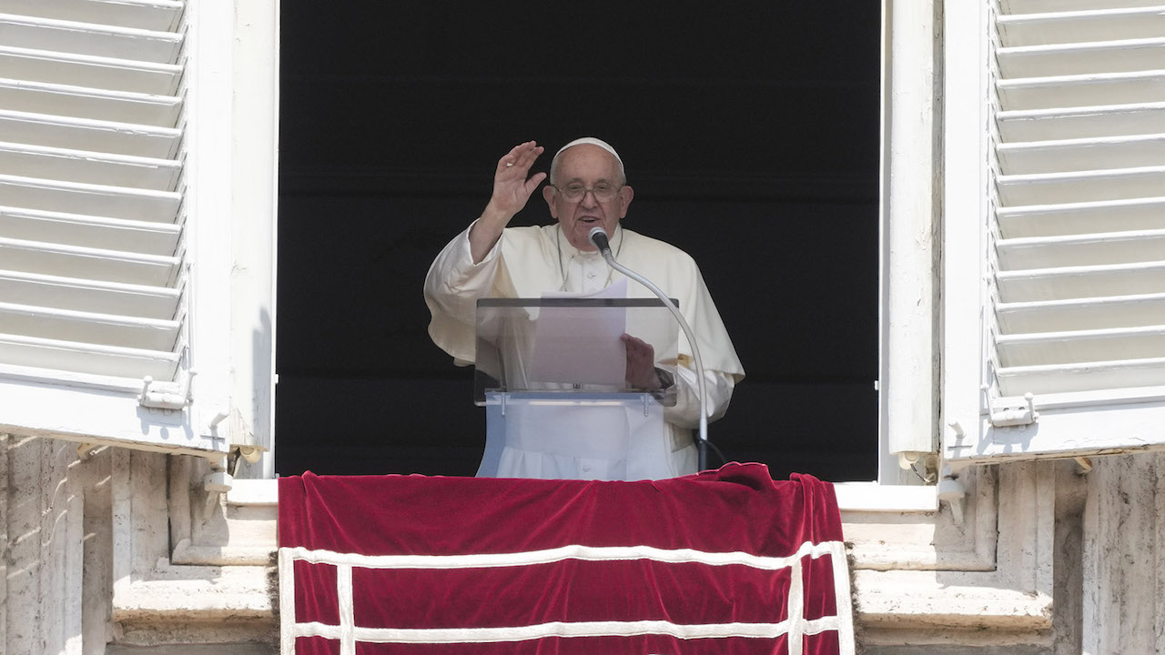 Pope Francis, dressed in white robes, recites a blessing out of a window with a red cloth.