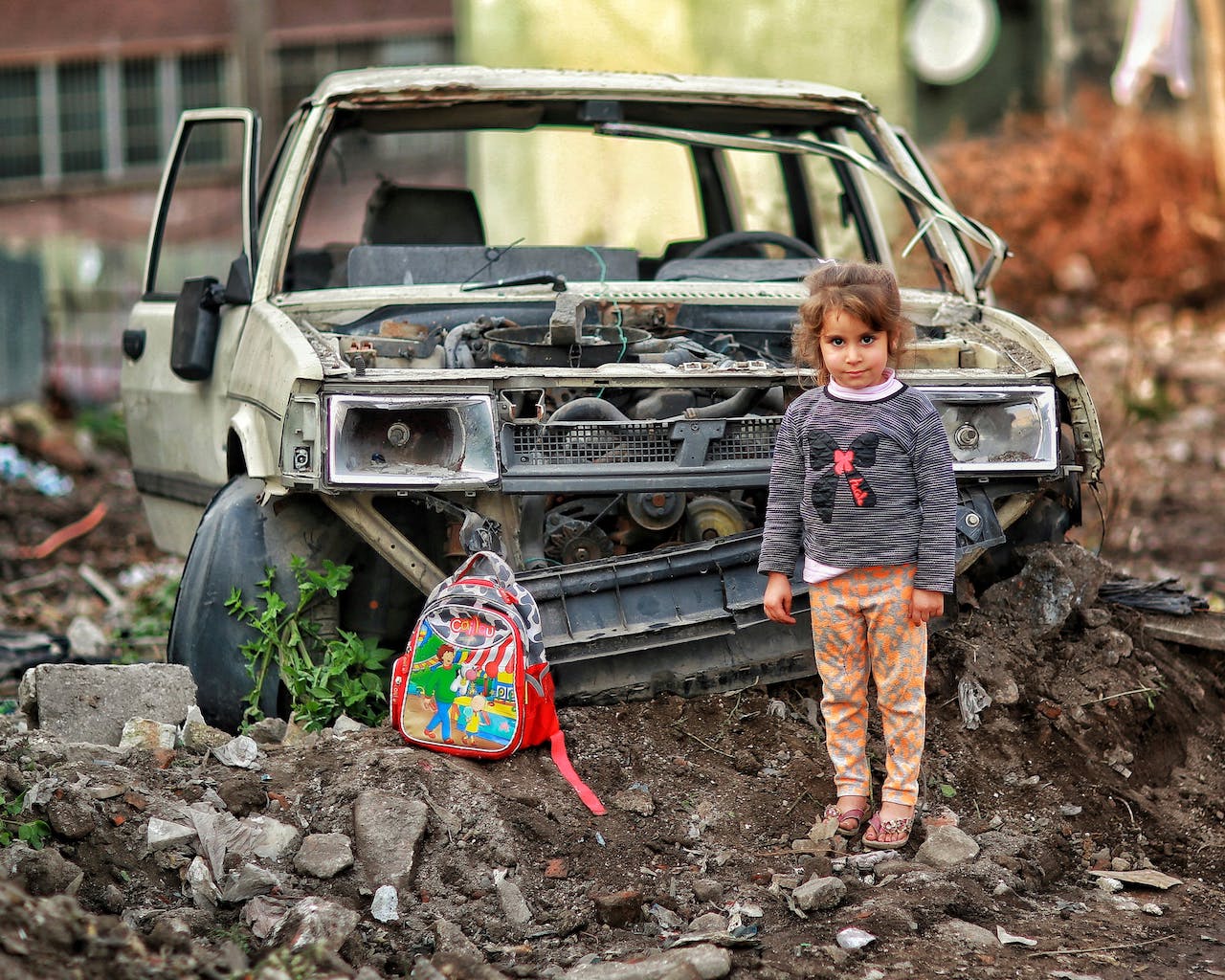 Girl with brightly colored backpack standing in front of car ruins