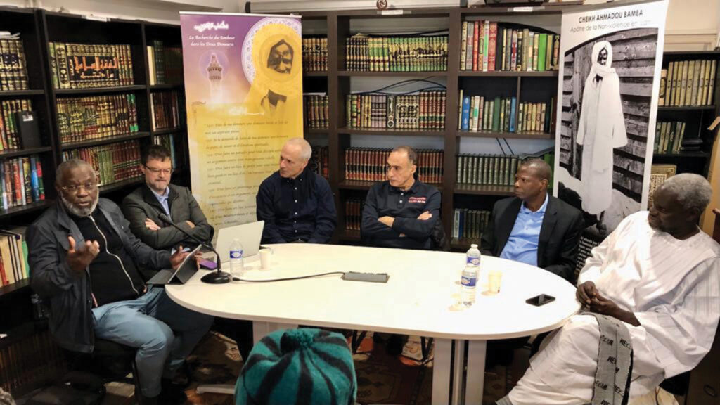 Jean-Claude Girondin, left, pastor of Villeneuve-le-Comte Mennonite Church near Paris, addresses the gathering at the Murid-Mennonite encounter. With him, from left, are Matthew Krabill, Jonathan Bornman, Abdullah Fahmi, Cheikh Babou and Mortalla Mboup. The background posters feature the Murids’ founder, Shaykh Amadu Bamba Mbakke. — Andres Prins