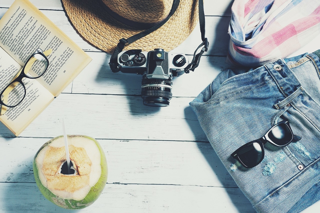 straw hat, camera, sunglasses, jeans and a coconut with a straw in it, against a white boardwalk background
