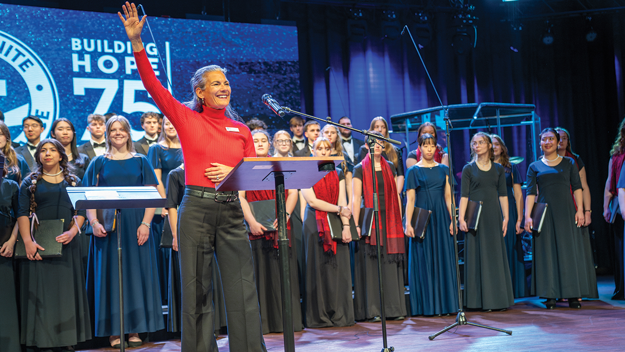 Lana Tieszen, MDS Region 3 vice chair, greets the celebration worship service crowd, backed by the Bethel, Hesston and Tabor college choirs. — Paul Hunt/MDS