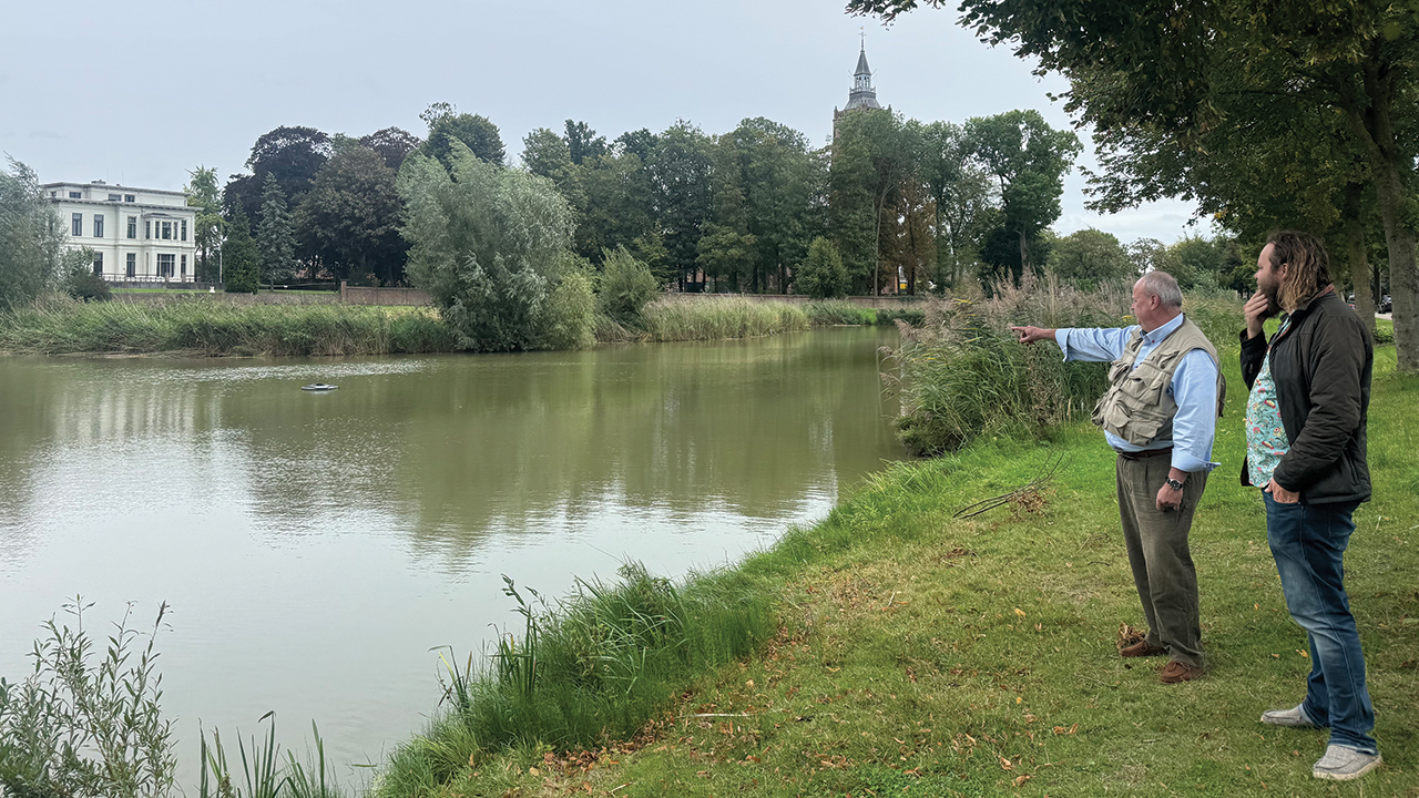Theo Witjes, chair of the Historical Society of Old Asperen, points out possible locations for a statue of Anabaptist martyr Dirk Willems in the former moat that froze over in 1569, enabling Willems’ escape. At right is Witjes’ son and fellow historical society member Freek Witjes. Asperen’s Dutch Reformed Church — a Catholic church when it served as Willems’ prison after his recapture — stands in the background. — Historical Society of Old Asperen