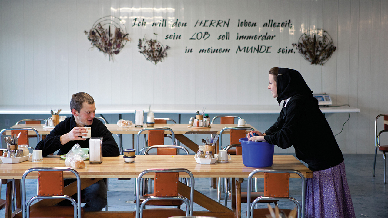 Late to lunch after chores in the dairy barn, Josh Wurtz catches up with Chantel Hofer as she wipes tables in the Deerboine Colony dining room near Alexander, Man., in 2010. The text on the wall comes from Psalm 34:1, “I will bless the Lord at all times: his praise shall continually be in my mouth.” — Tim Smith