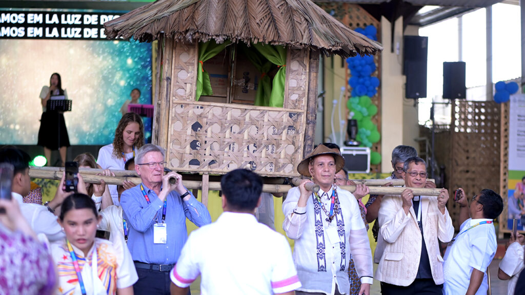 From left, Mennonite World Conference President Henk Stenvers, Lumban Mennonite Bible Church resident pastor Richard Rancap and Integrated Mennonite Church President Eladio Mondez carry a traditional nipa hut to represent the Filipino value of Bayanihan, in which neighbors work together to move a home in times of need. Rancap is also head of IMC’s mission department. — RG Photography/MWC