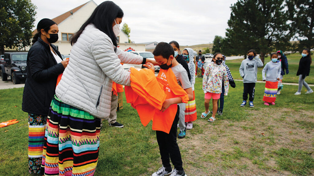 Students at Red Cloud Indian School wait in line to receive orange T-shirts after an assembly Sept. 30, 2021, in Pine Ridge, S.D. Students and teachers wore orange in solidarity with Indigenous children of past generations who suffered cultural loss, family separation and sometimes abuse and neglect while compelled to attend hundreds of residential schools that once dotted the map across the United States. — Emily Leshner/AP